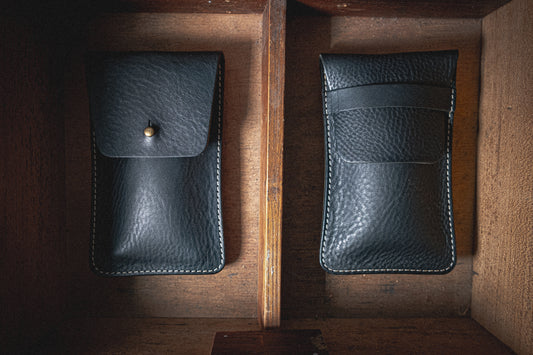 Two cases from the "Earth tones" Pebbled Veg-Tan collection are placed vertically in a wooden compartment. The left black leather case features a flap with a button closure, while the right one has an open top design with a small stitched pocket on the front. Both cases boast a textured finish and white stitching along the edges.