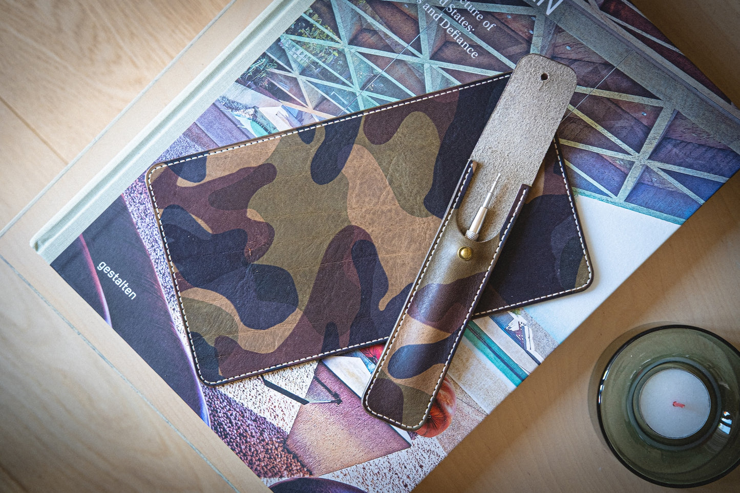 The watchpouch kit - PACKAGEDEAL, featuring a veg-tan leather camouflage-patterned wallet and matching strap, lies on a stack of architecture book covers. A nearby tealight candle in a glass holder enhances the warmth of the wooden surface with its glow.
