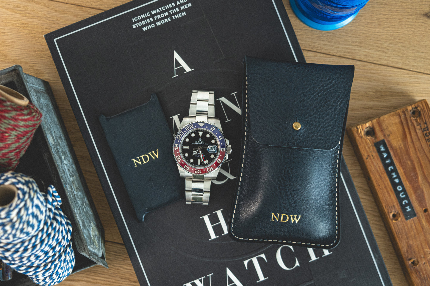 A silver wristwatch with a red and blue bezel is displayed on a book titled "A Man & His Watch." The watch comes with a black pouch and a black leather case, both inscribed with “NDW.” Partially visible on the sides are colored cords and a wooden box from the Pebbled veg-tan collection "Earth tones.