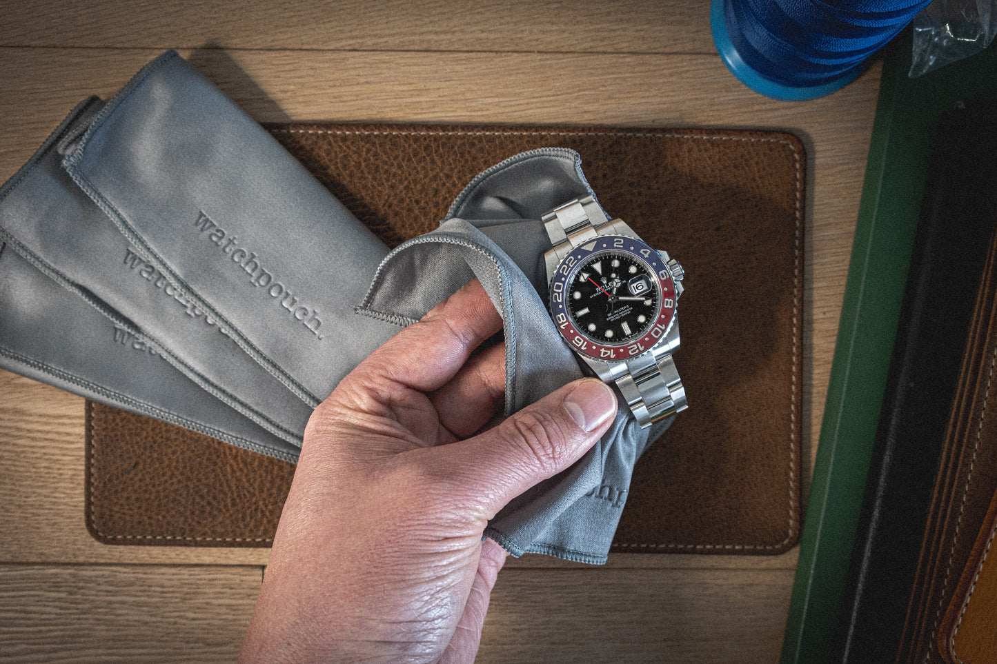 A hand holding a silver wristwatch with a black and red bezel, partially wrapped in a gray cloth. More gray clothes with "Watchbox" etched on them lie beneath the hand on top of a textured surface from the Pebbled veg-tan collection "Earth tones," and a blue container is visible in the upper right corner.