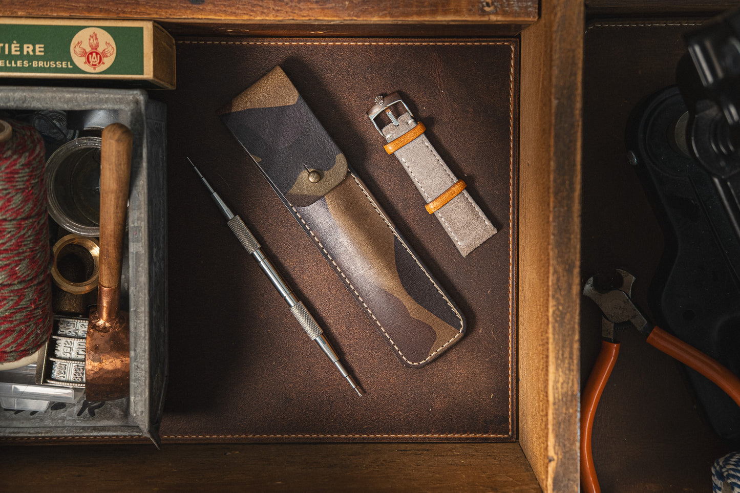 A neatly organized wooden drawer containing a Toolpouch for springbar-tool, a leather watch strap, a matching leather case, a spool of thread, a box of matches, and a small pair of pliers. The items are arranged against a dark brown background with precision for optimal watch care.