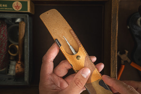 A person holds a Toolpouch for springbar-tool with one slot open, revealing a precision screwdriver. The surrounding workspace includes various tools such as a hammer, a pair of pliers, and a wooden-handled tool—ideal for both general tasks and specialized watch care.