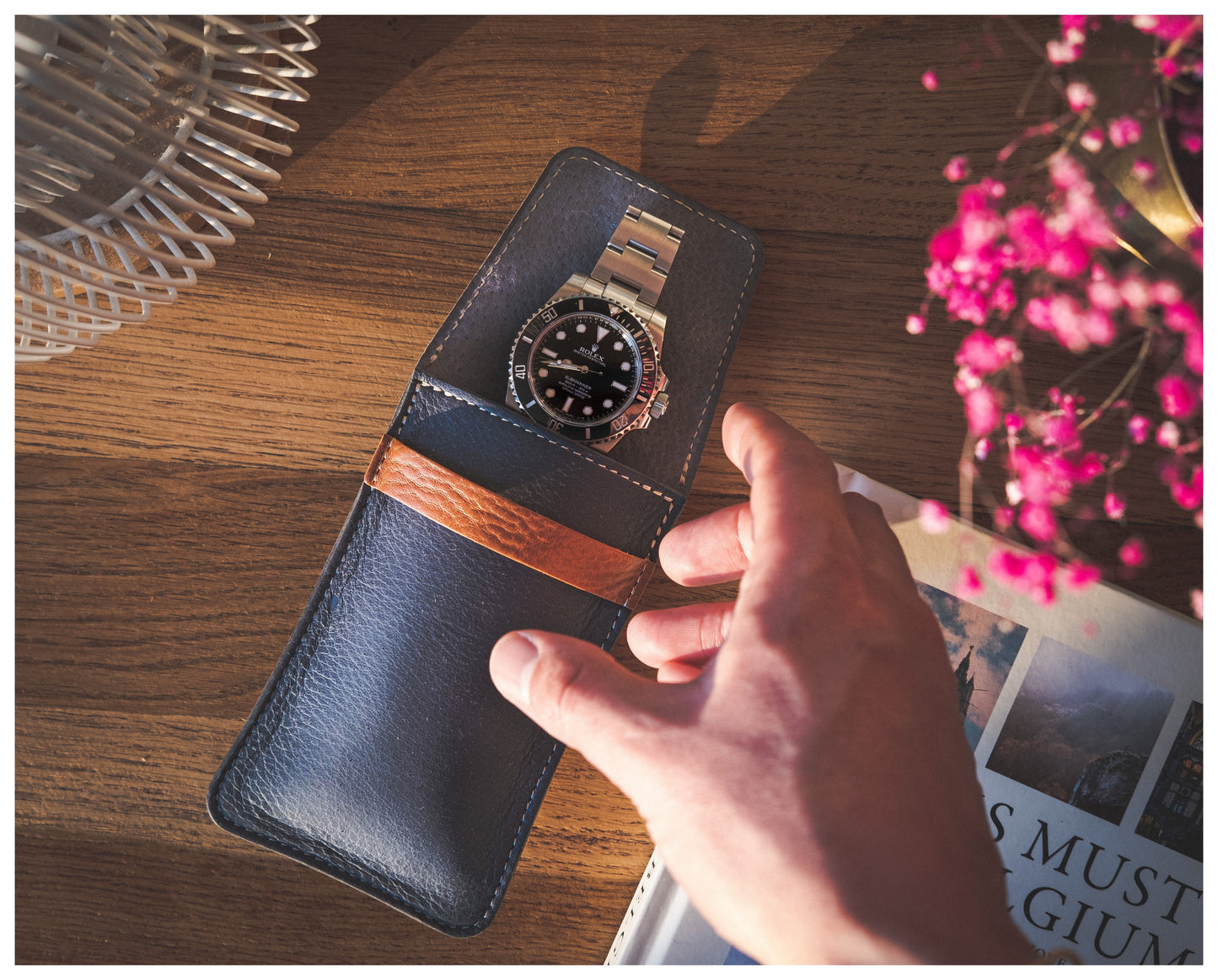 A hand reaches for a silver watch nestled in a pouch from the Soft Layered Premium Pouch Collection (with lining), handmade in Antwerp, resting on the wooden table. Nearby, a book with visible pages and a vase with pink flowers add to the warmly lit scene.