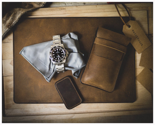 A luxury stainless steel watch with a black dial is displayed on a gray cleaning cloth atop a leather mat. Next to it is a patina leather wallet, a vintage brown watchpouch, and a tag labeled "watchout." The scene exudes elegance and sophistication.