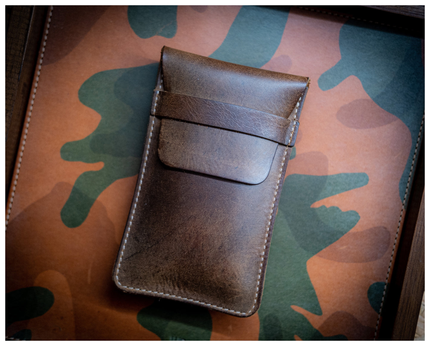 A vintage brown phone case with a flap cover rests on a surface with a camouflage pattern. The case, made of patina leather, showcases a smooth, slightly worn texture and detailed stitching along the edges, exuding an elegant yet rugged charm reminiscent of a classic watchpouch.