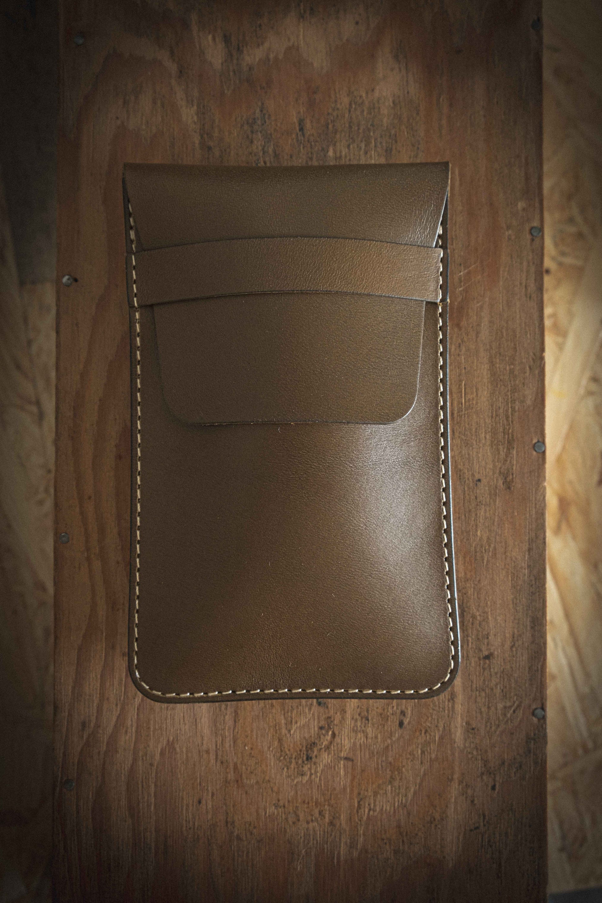 An olive leather pouch crafted from veg tan leather with white stitching is displayed on a textured wooden surface. The pouch, part of our SAMPLE SALE collection, features a flap closure and offers simple elegance with personalization available for a unique touch.