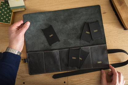 A person is holding an open leather folder from the Watchroll collection, available in various colors, featuring "D&G" embossed on several small black envelopes inside. The leather folder includes multiple pockets and an attached strap, designed to hold personalized watchrolls. The scene is set on a wooden surface.