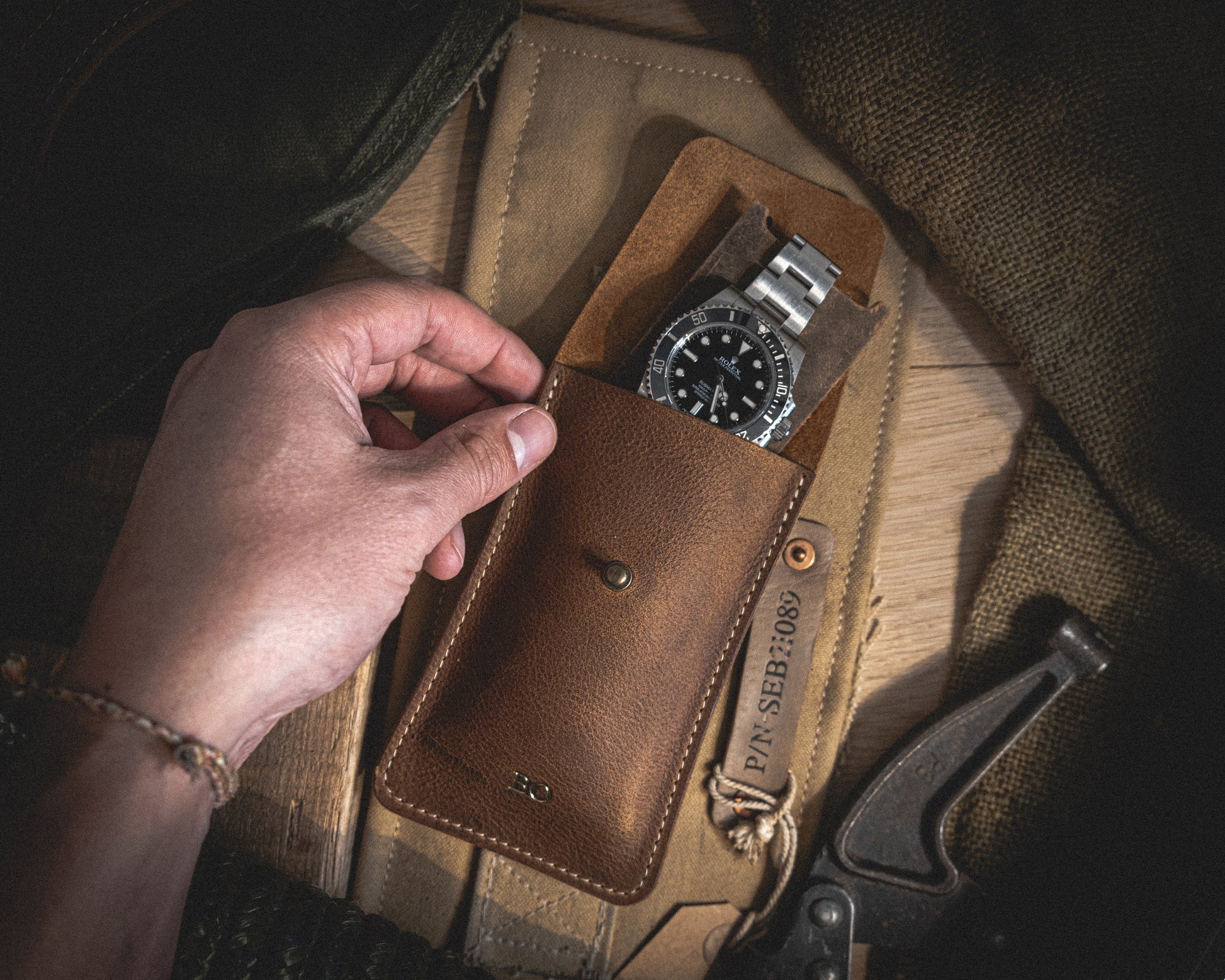 A hand reaches for a silver and black diving watch stored in a Rugged chestnut, a single watch pouch made of patina-loving, vegetable-tanned leather. Resting on a light brown suede mat and surrounded by various tools and fabrics, the scene exudes a rustic ambiance with subdued lighting.