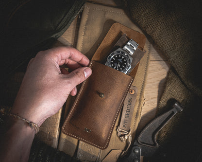 A hand reaches for a silver and black diving watch stored in a Rugged chestnut, a single watch pouch made of patina-loving, vegetable-tanned leather. Resting on a light brown suede mat and surrounded by various tools and fabrics, the scene exudes a rustic ambiance with subdued lighting.