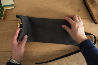 A person with a wristwatch on their left hand is unfurling a black leather pouch with a brass D-ring and strap on a wooden surface. Nearby, there's a green box, personalized watchrolls from the Watchroll collection in various colors, and a black brush, with a partially visible object in the top left corner.