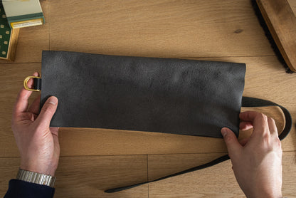 Two hands are holding a dark gray leather piece from the Watchroll collection (var. colors), featuring a brass ring on one end. A leather strap extends from the piece, making it ideal for securing watchrolls. The background showcases a wooden surface with visible edges of boxes and a brush, adding a personalized touch.