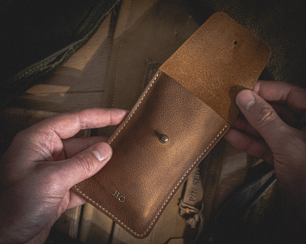 A person is holding an open Rugged chestnut leather pouch with visible stitches. The pouch, which appears to be a watch pouch, has a button closure and the initials "B.O" embossed on the lower front. The background consists of a wooden surface and another leather item, partially visible.