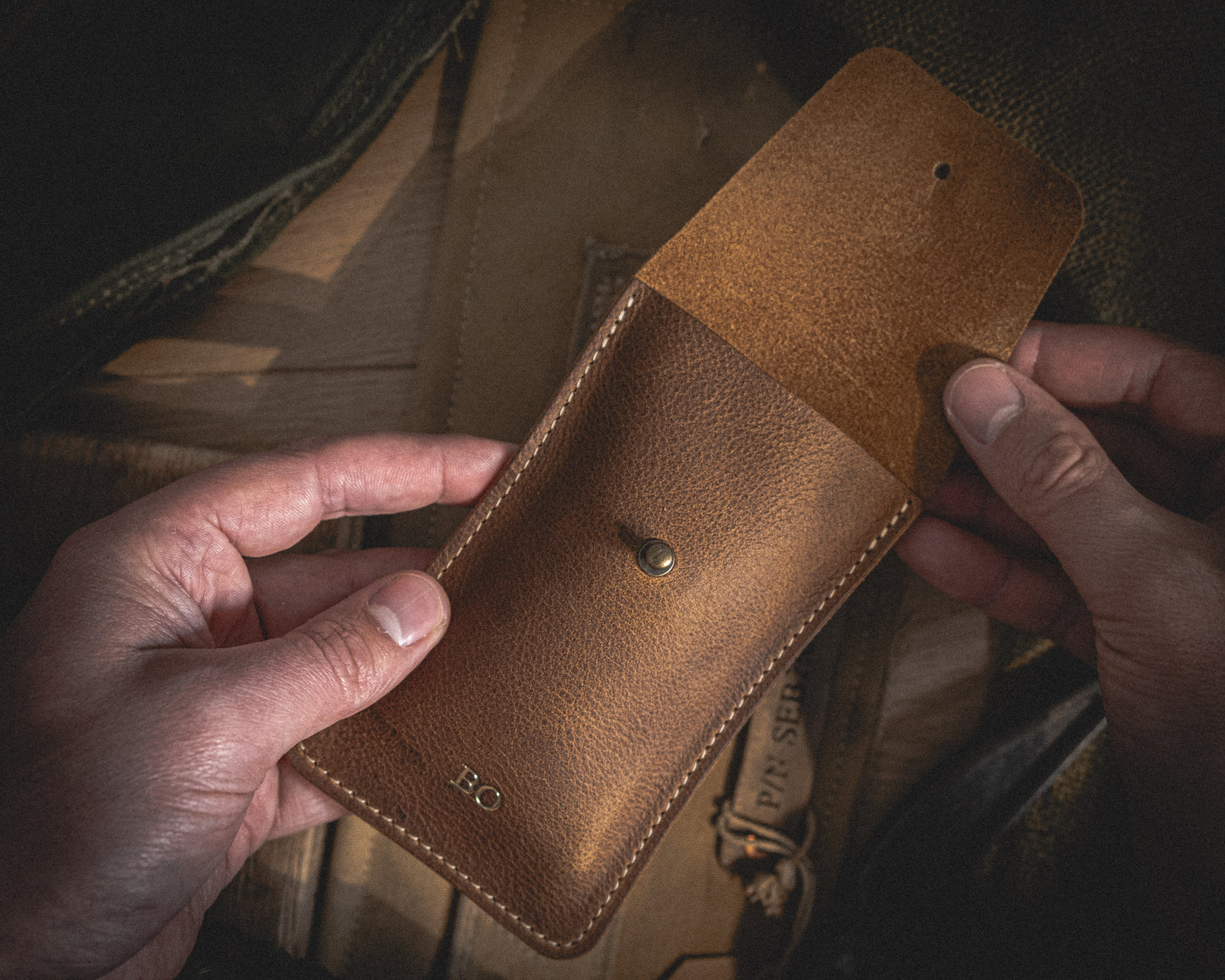 A person is holding an open Rugged chestnut leather pouch with visible stitches. The pouch, which appears to be a watch pouch, has a button closure and the initials "B.O" embossed on the lower front. The background consists of a wooden surface and another leather item, partially visible.