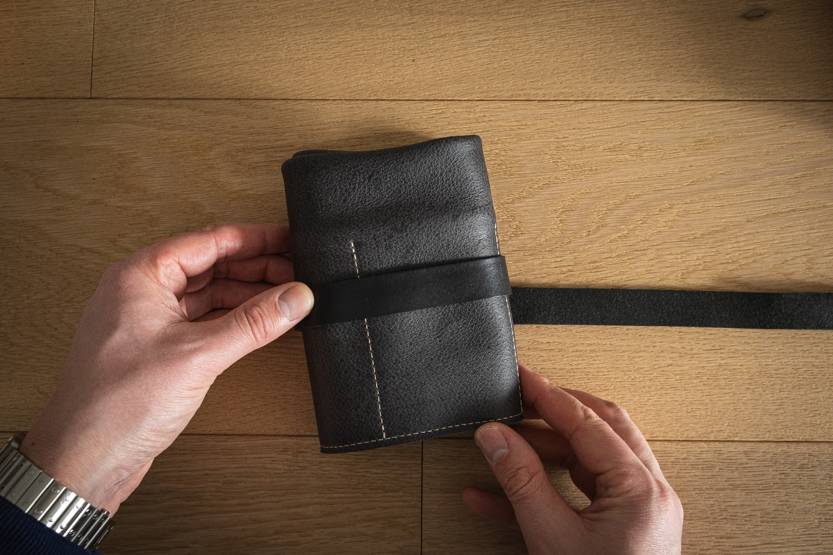 A pair of hands is holding a rolled-up black leather Watchroll from the Watchroll collection on a wooden surface, with its strap securing it tightly. The person is wearing a silver watch on their left wrist. The personalized leather pouch features visible stitching and boasts a smooth texture.