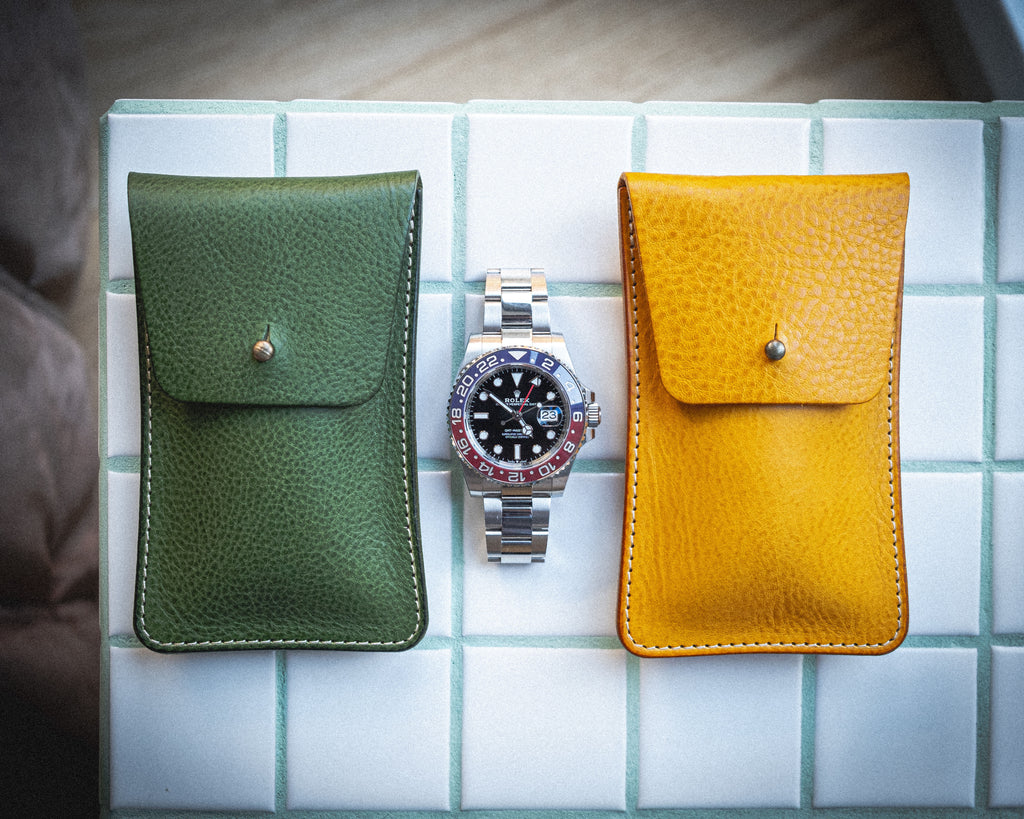 A silver wristwatch with a black dial and red accents is displayed between two leather pouches from the Pebbled veg-tan collection "vibrant", handmade in Antwerp. The green pouch is on the left, and the yellow pouch is on the right, both crafted from pebbled veg-tan leather and feature small button clasps. The items are arranged on a white, tiled surface.