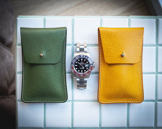 A silver wristwatch with a black dial and red accents is displayed between two leather pouches from the Pebbled veg-tan collection "vibrant", handmade in Antwerp. The green pouch is on the left, and the yellow pouch is on the right, both crafted from pebbled veg-tan leather and feature small button clasps. The items are arranged on a white, tiled surface.