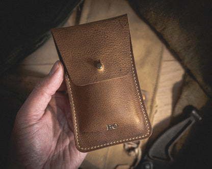 A hand holds the Rugged Chestnut leather pouch with a buttoned flap. The pouch features visible stitching and the initials "R.O." embossed near the bottom. In the background, there is a blurred view of various objects, including a piece of green fabric and a metallic tool.