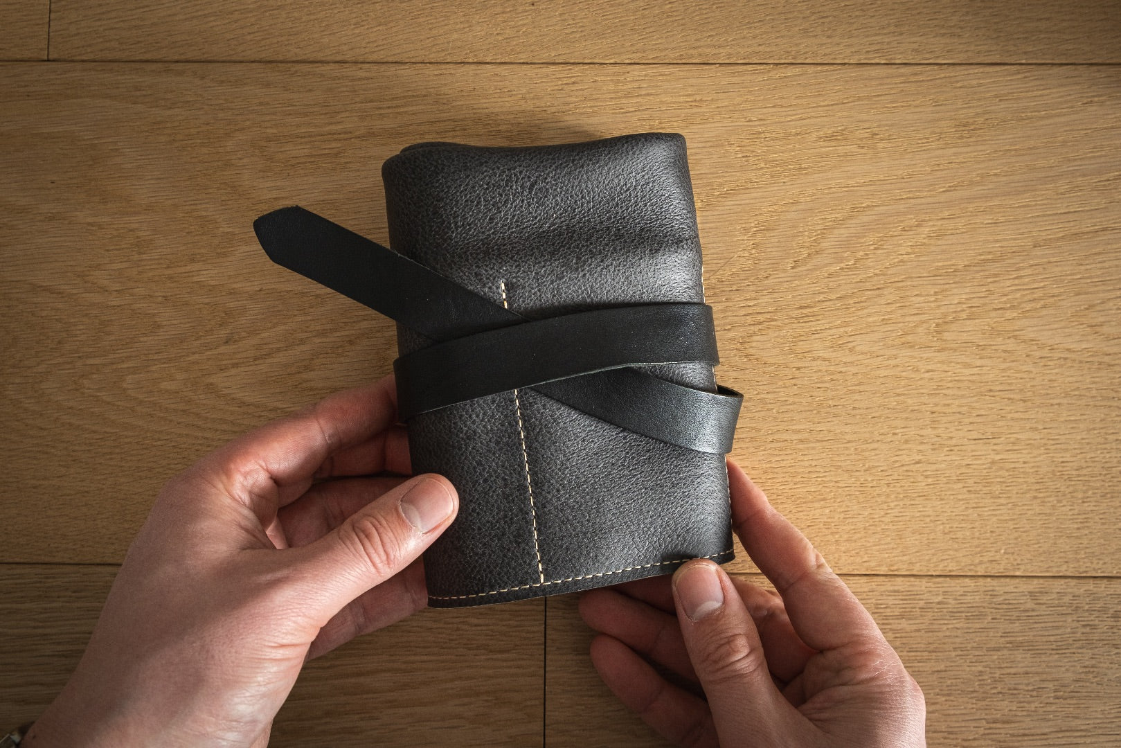 A pair of hands holding a rolled-up case from the Watchroll Collection, made of black leather and secured with a leather strap. The background is a light-colored wooden surface, ideal for showcasing personalized watchrolls.