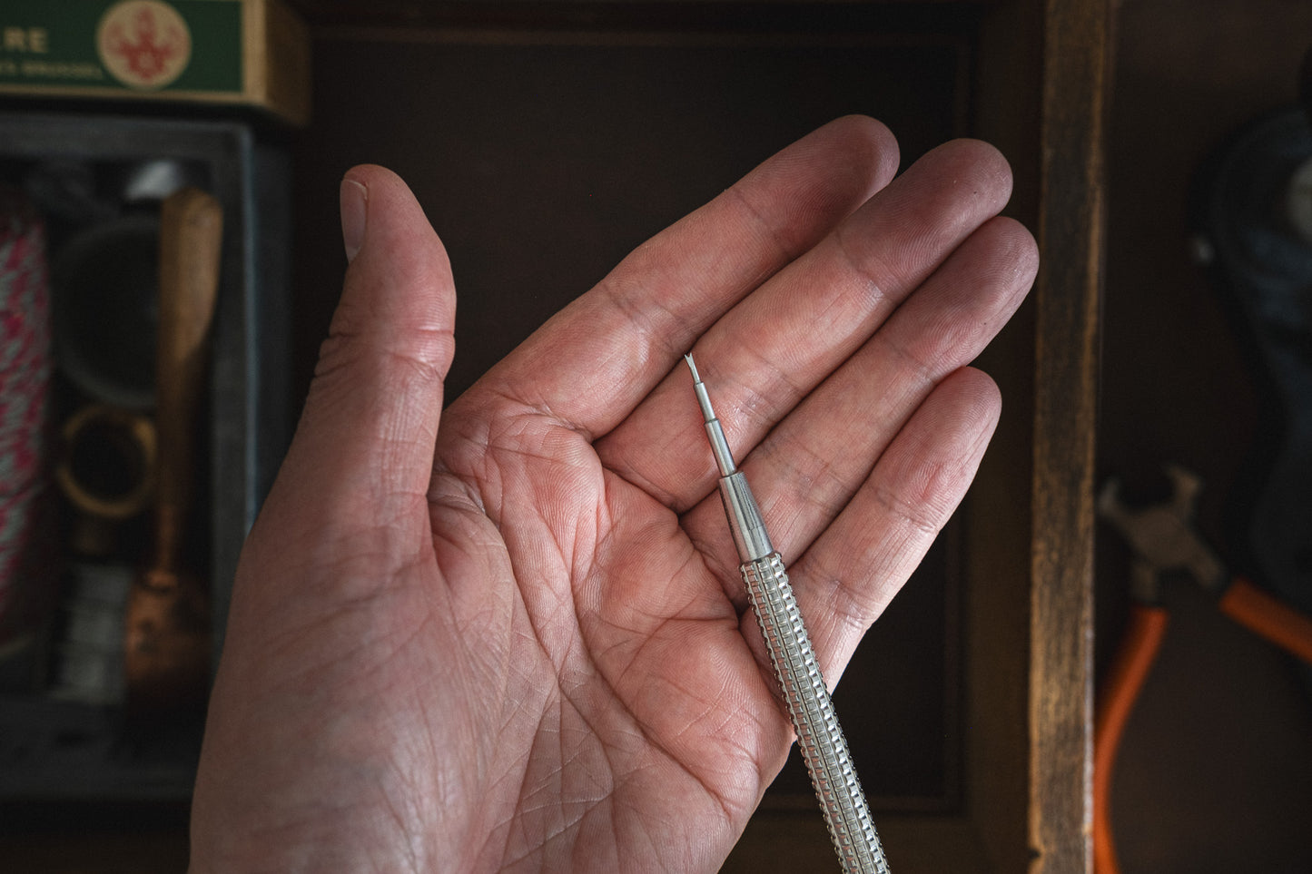 A hand holds a precision screwdriver featuring a slender, metallic handle and a small tip, ideal for tasks requiring fine adjustments. The background showcases leather tool pouches and the Toolpouch for springbar-tool, alongside a box containing various tools and materials.