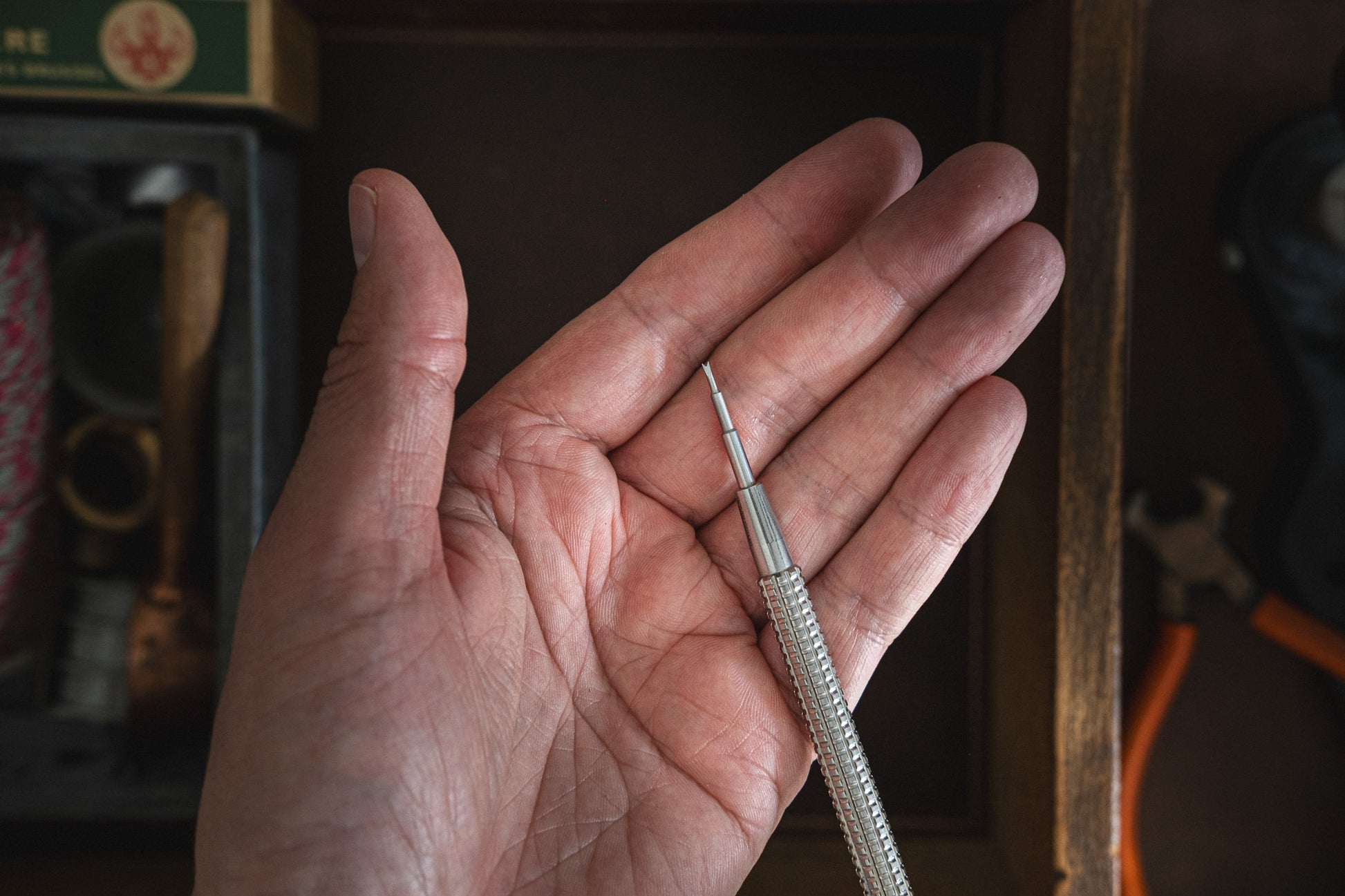 A hand holds a precision screwdriver featuring a slender, metallic handle and a small tip, ideal for tasks requiring fine adjustments. The background showcases leather tool pouches and the Toolpouch for springbar-tool, alongside a box containing various tools and materials.