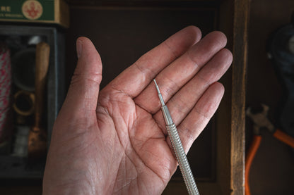 A hand holds a precision screwdriver featuring a slender, metallic handle and a small tip, ideal for tasks requiring fine adjustments. The background showcases leather tool pouches and the Toolpouch for springbar-tool, alongside a box containing various tools and materials.