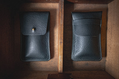 Two cases from the "Earth tones" Pebbled Veg-Tan collection are placed vertically in a wooden compartment. The left black leather case features a flap with a button closure, while the right one has an open top design with a small stitched pocket on the front. Both cases boast a textured finish and white stitching along the edges.