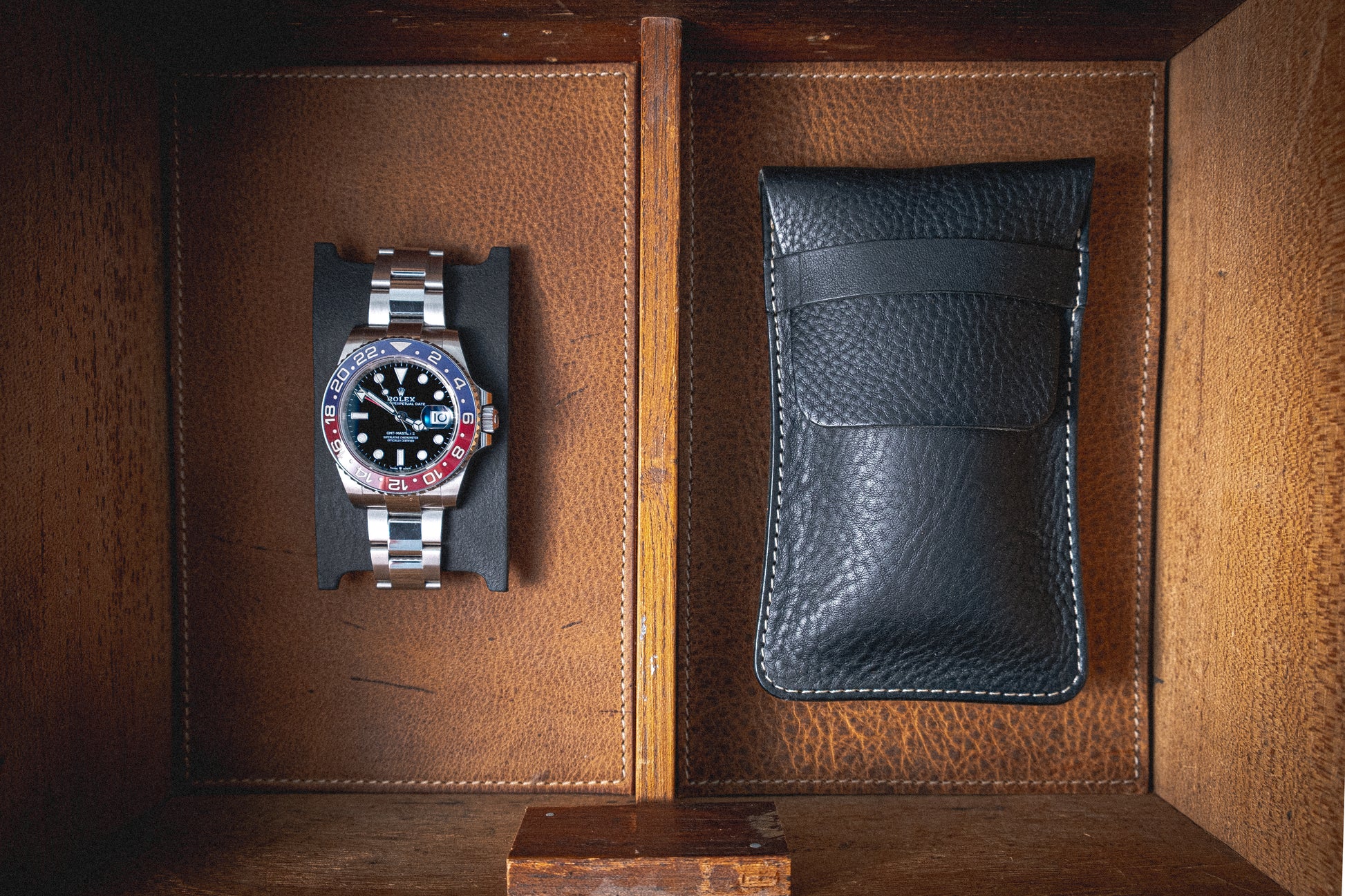 The Pebbled veg-tan collection "Earth tones" features a metal wristwatch with a blue and red bezel and a black watch face, elegantly presented in a rustic wooden box. The box, divided into two sections, houses the watch alongside a black leather pouch crafted from veg-tan leather, creating an inviting blend of earth tones perfect for personalization.