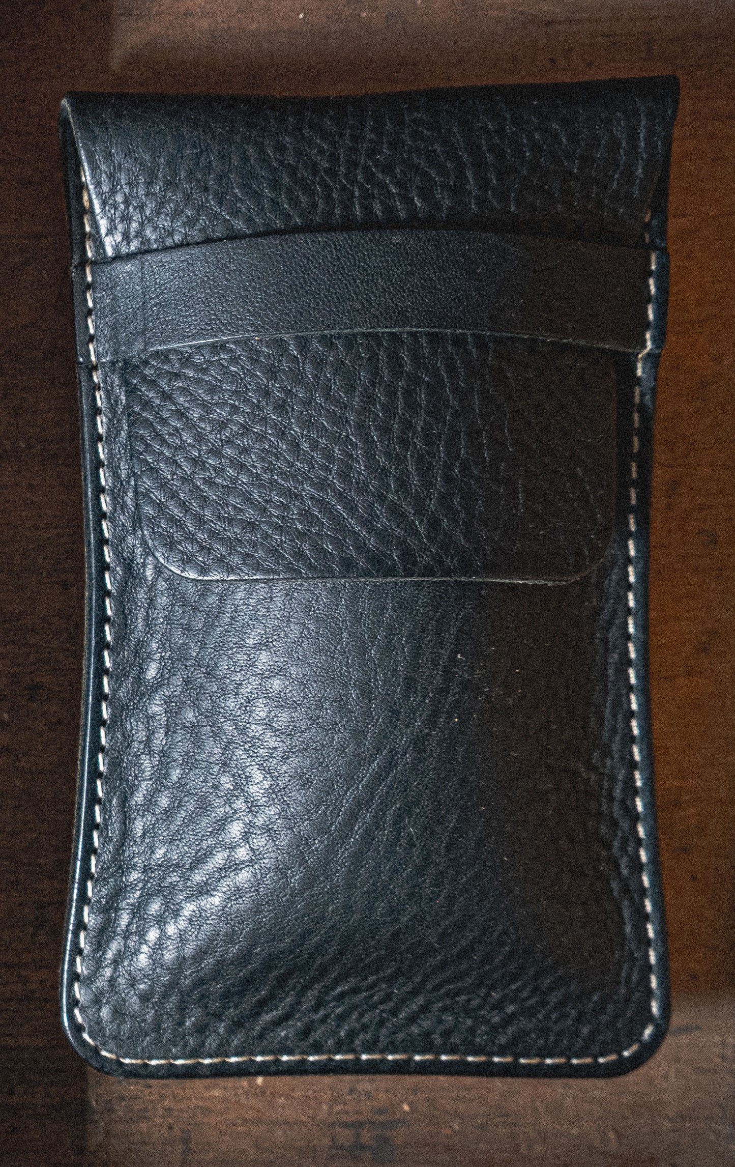 A black leather pouch from the Pebbled veg-tan collection "Earth tones" with a textured surface and white stitching around the edges lies on a wooden surface, perfectly complementing earth tones. The pouch boasts a front pocket and sleek design, offering personalization options for added uniqueness.