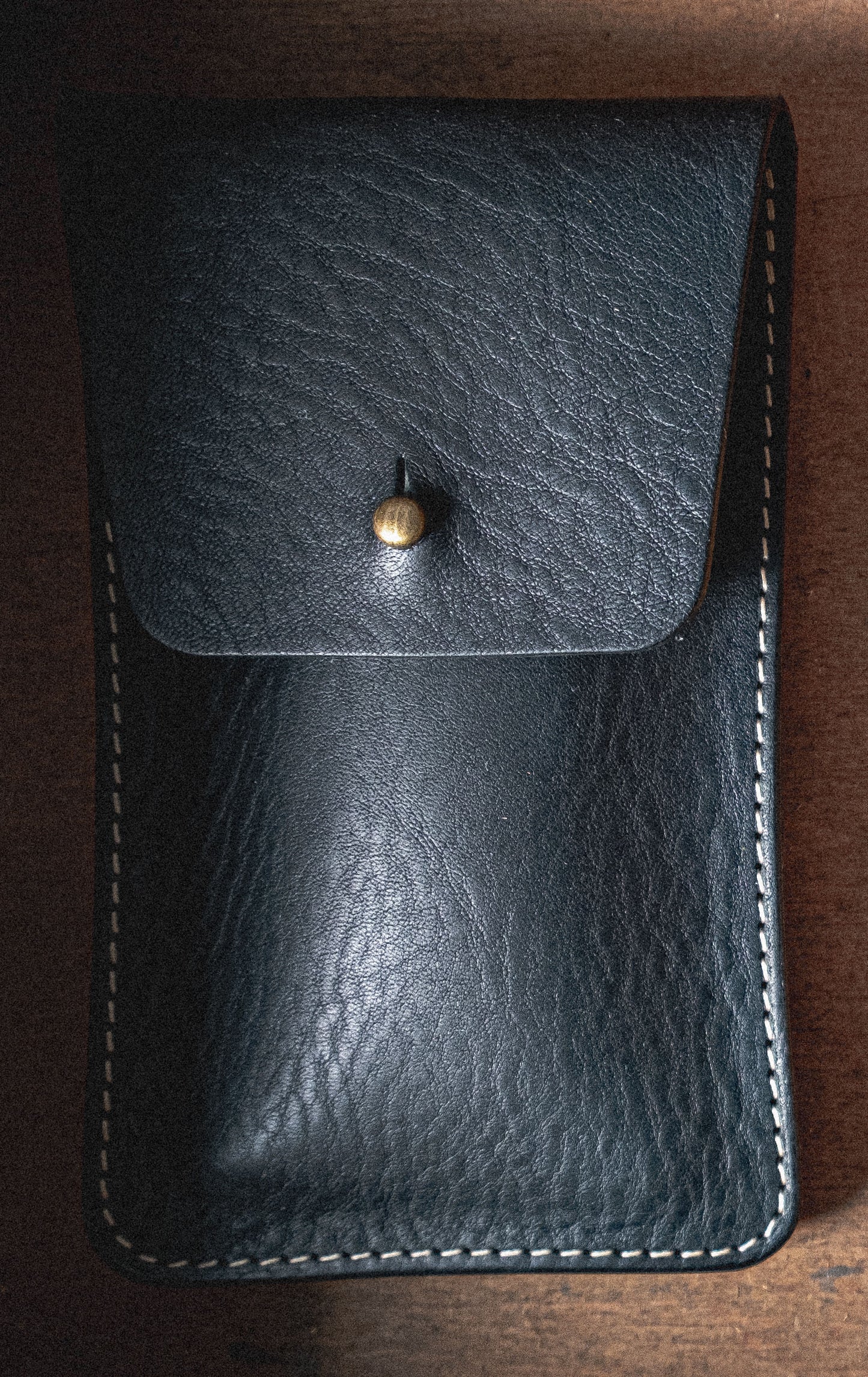 Introducing the Pebbled Veg-Tan Collection "Earth Tones": a black leather pouch with a textured surface, secured by a brass button. Crafted from veg-tan leather, this pouch features white stitching along the edges and is set against a wooden background. The earthy tones complement its rugged yet refined design, making it ideal for personalization.