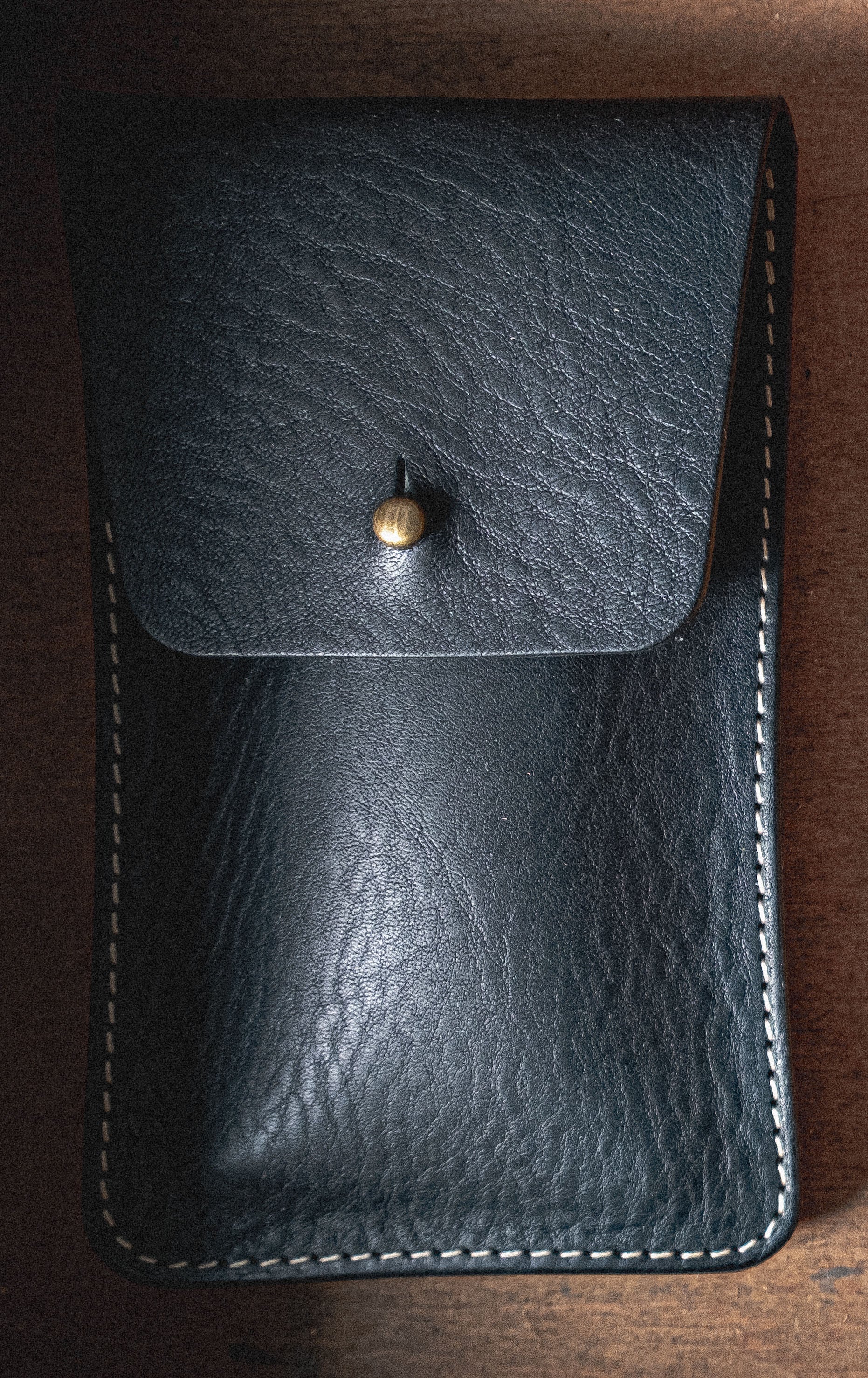Introducing the Pebbled Veg-Tan Collection "Earth Tones": a black leather pouch with a textured surface, secured by a brass button. Crafted from veg-tan leather, this pouch features white stitching along the edges and is set against a wooden background. The earthy tones complement its rugged yet refined design, making it ideal for personalization.