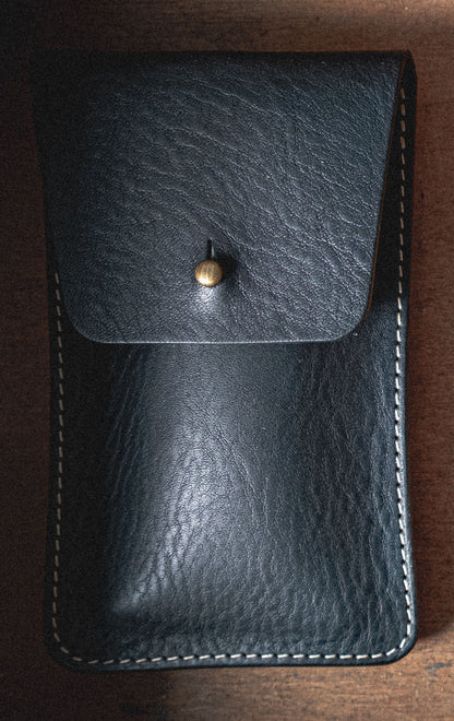 Introducing the Pebbled Veg-Tan Collection "Earth Tones": a black leather pouch with a textured surface, secured by a brass button. Crafted from veg-tan leather, this pouch features white stitching along the edges and is set against a wooden background. The earthy tones complement its rugged yet refined design, making it ideal for personalization.