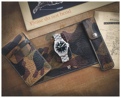 A Veg-tan CAMO COLLECTION leather watch roll with a stainless steel watch is placed on a light wood surface. Beside it are a map, a microfiber cleaning cloth, and an envelope with "Please do not bend" printed in red. Wrapped in adventure, the scene hints at journeys to come.
