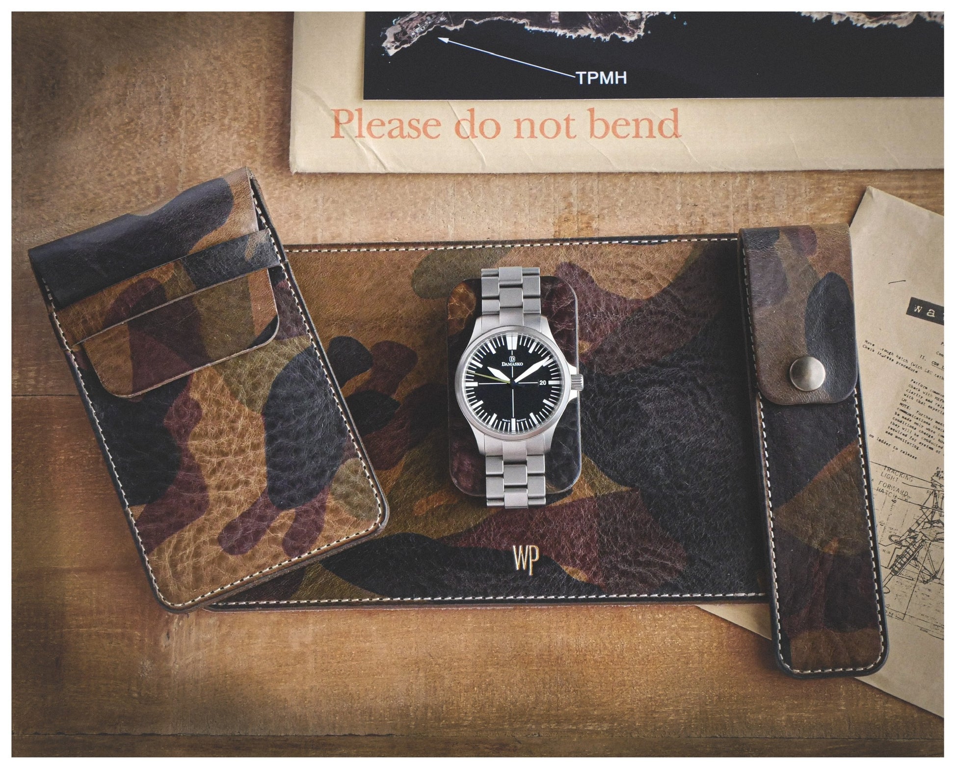 A watch from the Veg-tan CAMO COLLECTION featuring a silver wristwatch with a black dial and luminous markers is displayed on a camouflage leather mat. Surrounding it are two handmade watch pouches and a brown envelope labeled "Please do not bend" in red text. A map is partially visible on the right side.