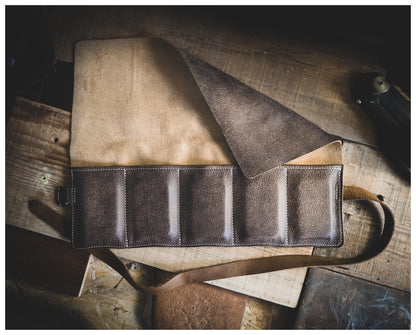 A versatile piece from the Watchroll Collection (available in various colors) is laid flat on a wooden surface. Partially open, it reveals a soft, suede-like interior. The leather strap and detailed stitching showcase its craftsmanship. Though designed as a rustic leather tool roll with multiple pockets, it also serves exceptionally well as one of the finest personalized watchrolls for travel.
