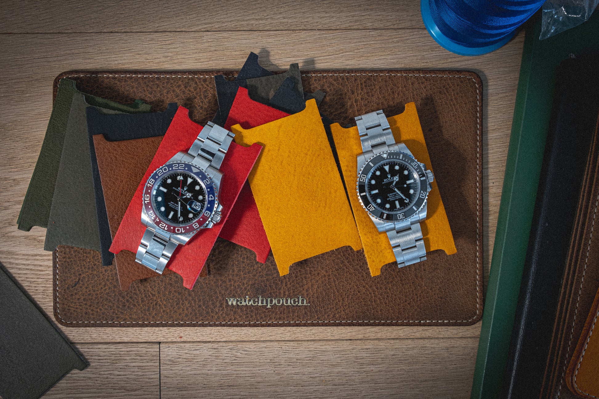 Two stainless steel watches with black faces on colored fabric swatches lie on a Soft Layered Premium Pouch Collection (with lining) labeled "watchpouch." Handmade in Antwerp, this premium watch accessory rests on a wooden surface adorned with various items, including a watch case and part of a blue water bottle.