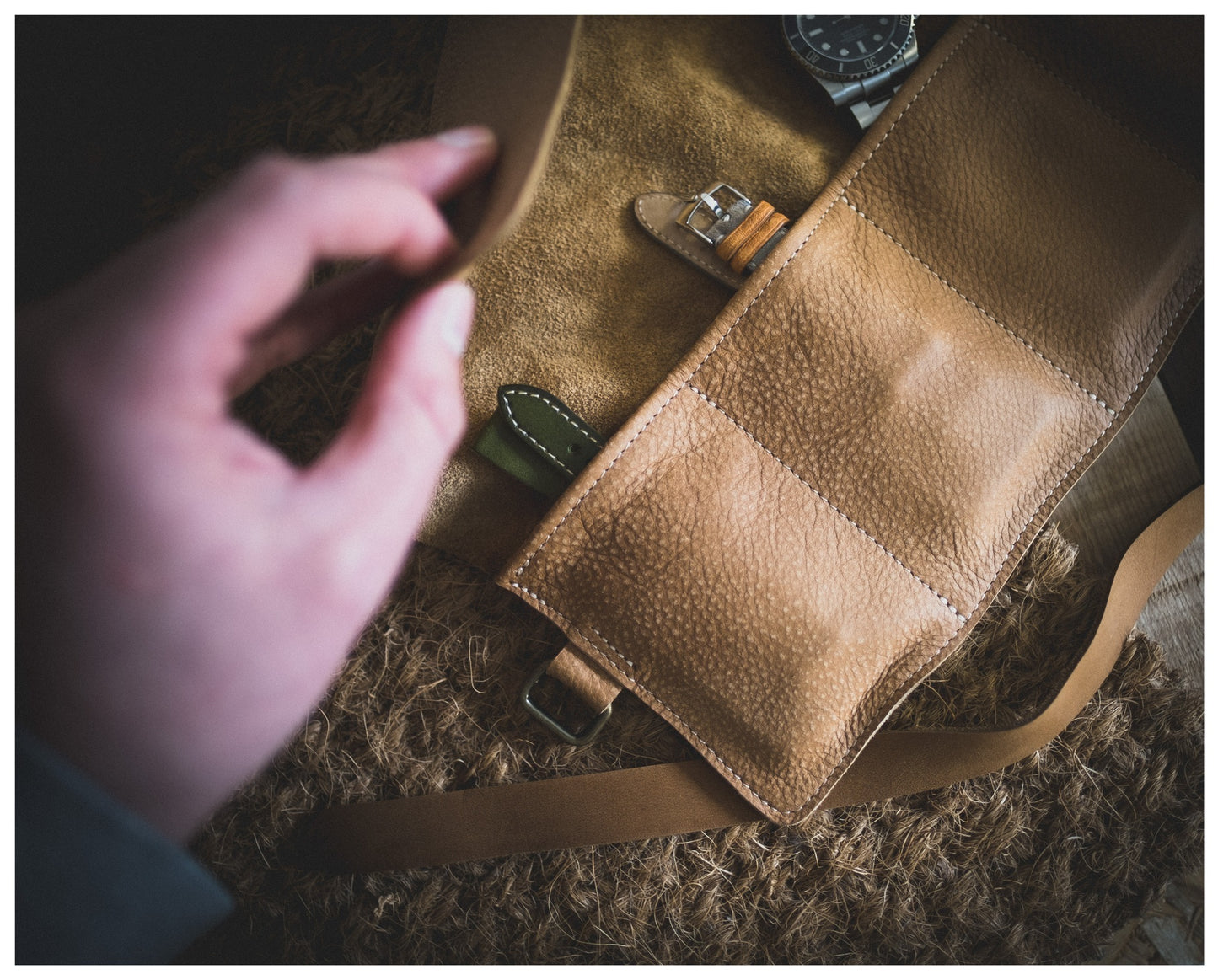 A hand unveils a personalized tan leather case from the Watchroll collection, revealing multiple compartments with one housing two watches. The textured leather case lies on a matching surface, showcasing one watch with a leather strap and its face partially visible.