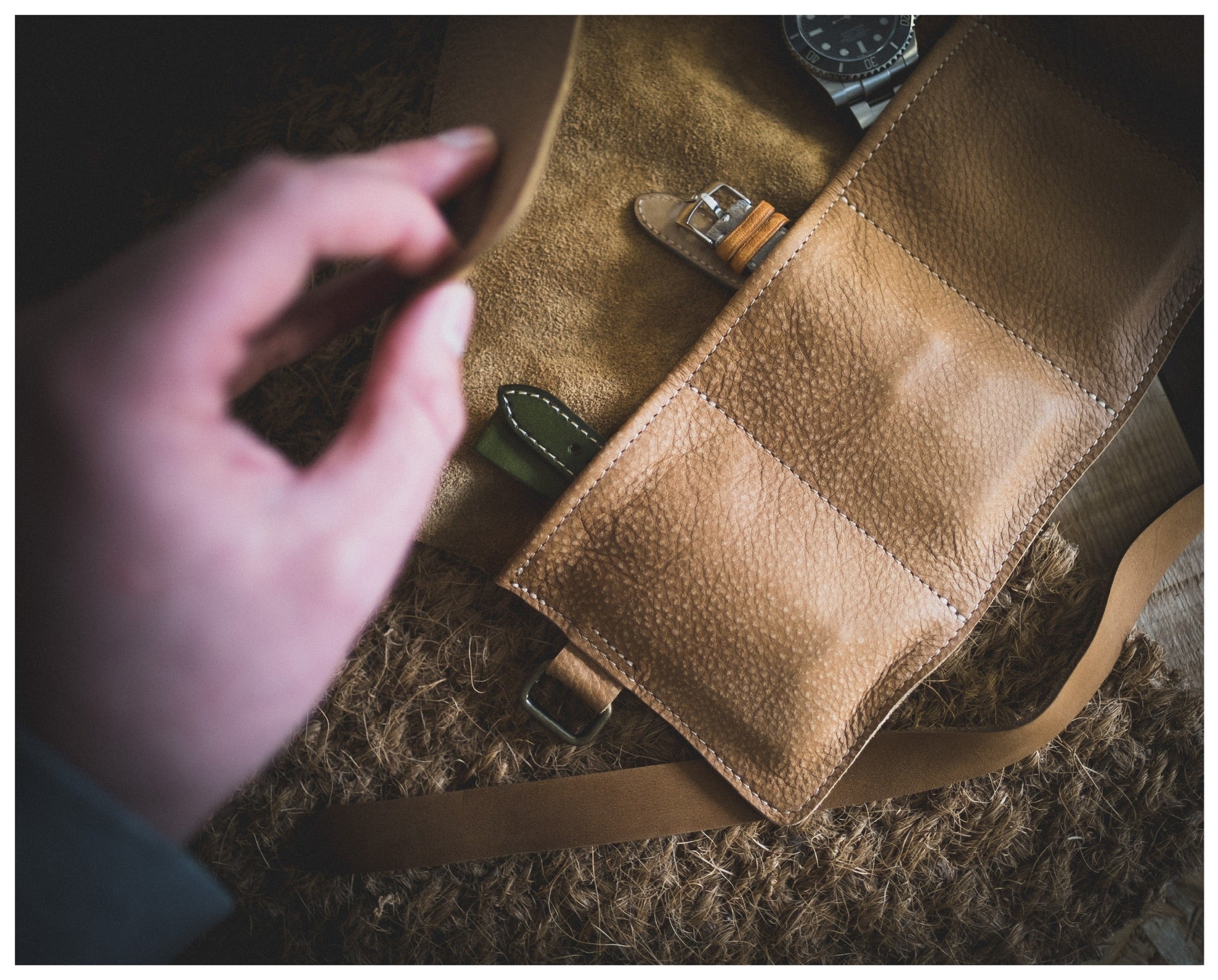 A hand unveils a personalized tan leather case from the Watchroll collection, revealing multiple compartments with one housing two watches. The textured leather case lies on a matching surface, showcasing one watch with a leather strap and its face partially visible.