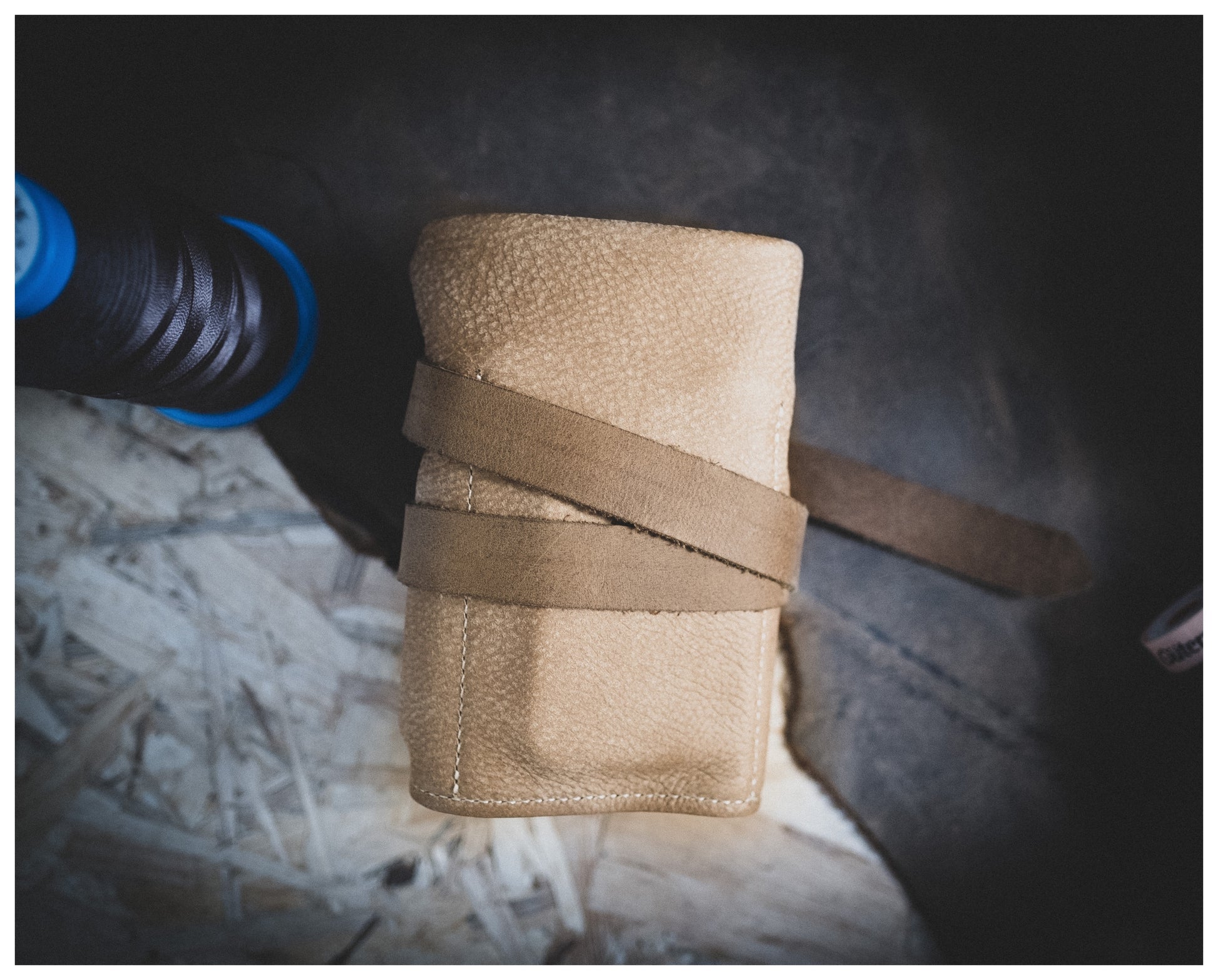 A small, folded leather pouch from the Watchroll collection lies on a wooden surface, secured with a strap wrapped around it. The textured leather adds to its charm. To the left of the pouch, there is a spool of dark thread. This personalized setup exudes a rustic and handmade feel.