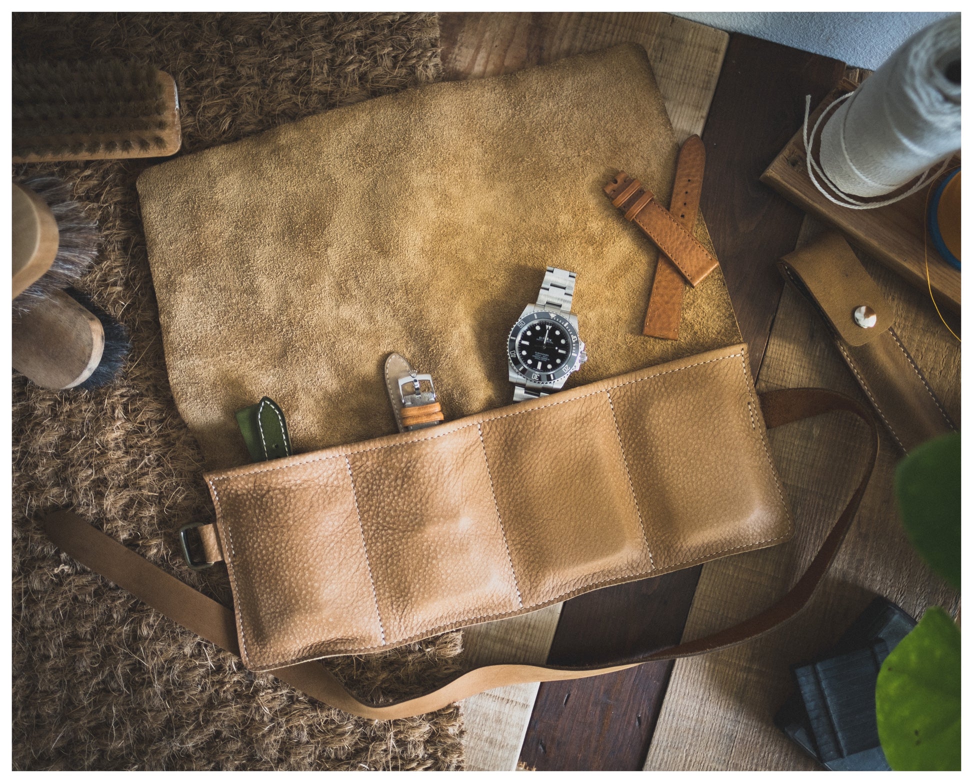 The tan leather piece from the Watchroll collection is open on a textured surface, showcasing a silver wristwatch and two extra watch straps. Surrounding it are various watch accessories and tools, including a brush and spools of thread. The scene emanates a vintage, handcrafted aesthetic.