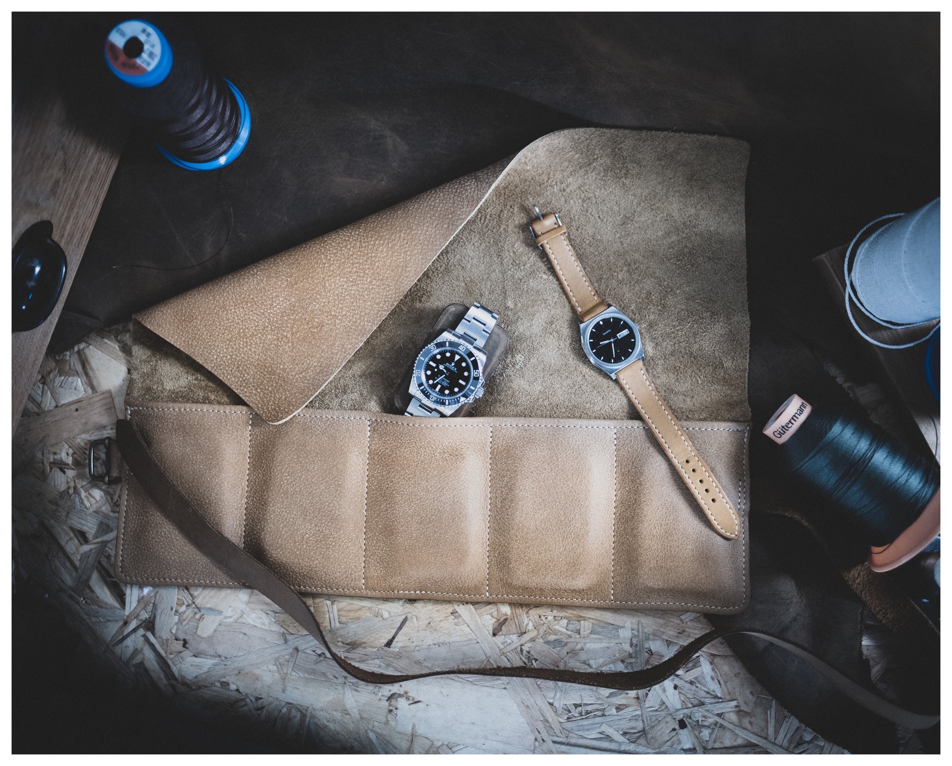 A personalized brown leather item from the Watchroll collection (available in various colors) is showcased on a wooden surface. It features several compartments and is accompanied by two wristwatches: one with a metal band and black dial, the other with a brown leather strap and black dial. Various sewing materials and threads are scattered around.