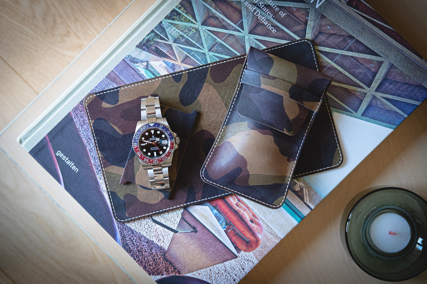 The watchpouch kit - PACKAGEDEAL features a camouflage watch holder and cardholder made from veg-tan leather on an architectural book cover. A luxury watch with a red and blue bezel is displayed, while a personalized glass candle holder adds elegance to the setup with earthy tones.