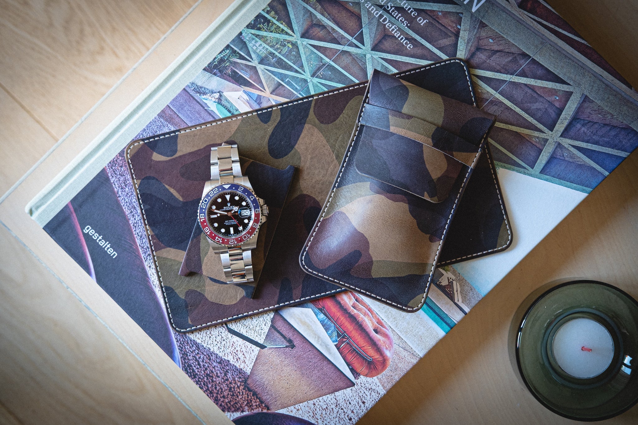 The watchpouch kit - PACKAGEDEAL features a camouflage watch holder and cardholder made from veg-tan leather on an architectural book cover. A luxury watch with a red and blue bezel is displayed, while a personalized glass candle holder adds elegance to the setup with earthy tones.