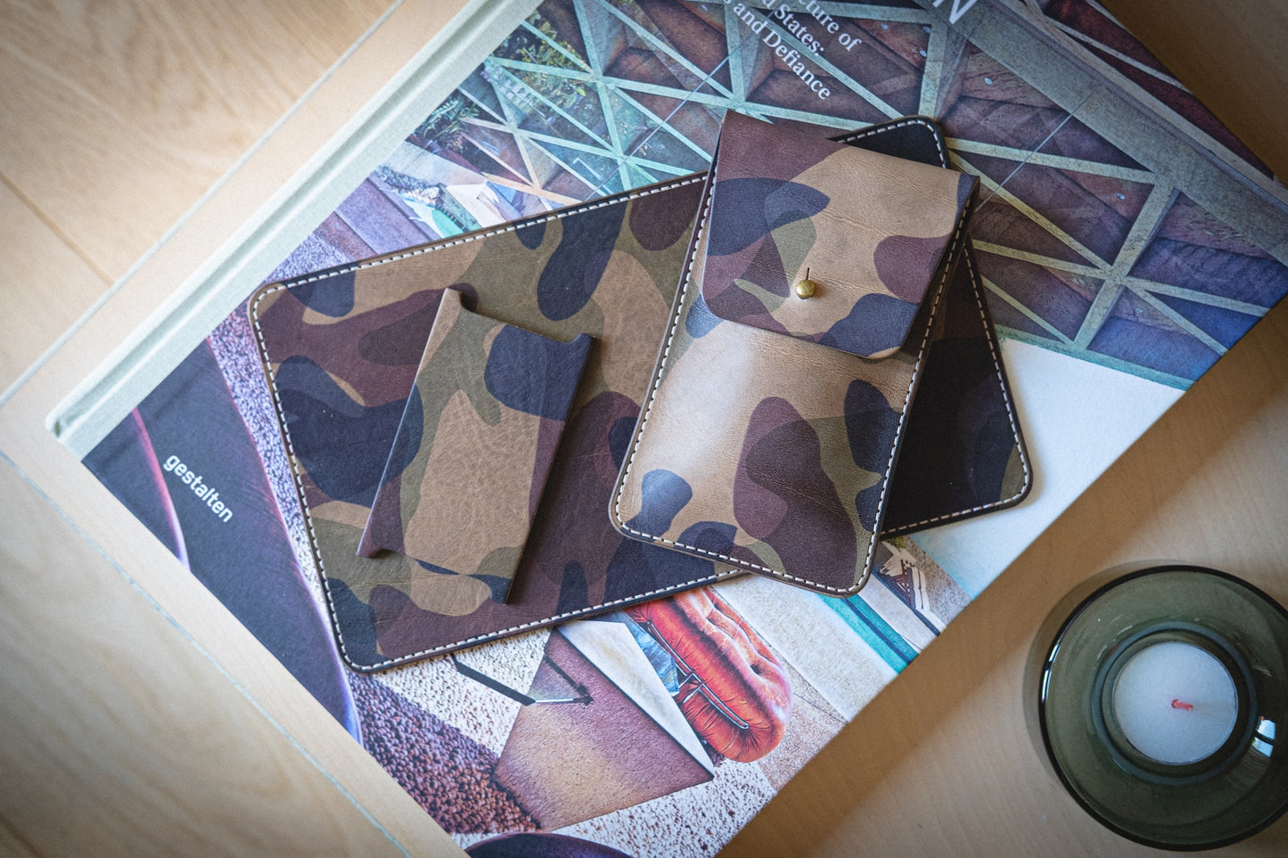 The watchpouch kit - PACKAGEDEAL, made from veg-tan camouflage-patterned leather, is displayed on an open book with architectural images. Nearby, a lit white candle in a glass holder adds warmth to the earth-toned wooden setting.