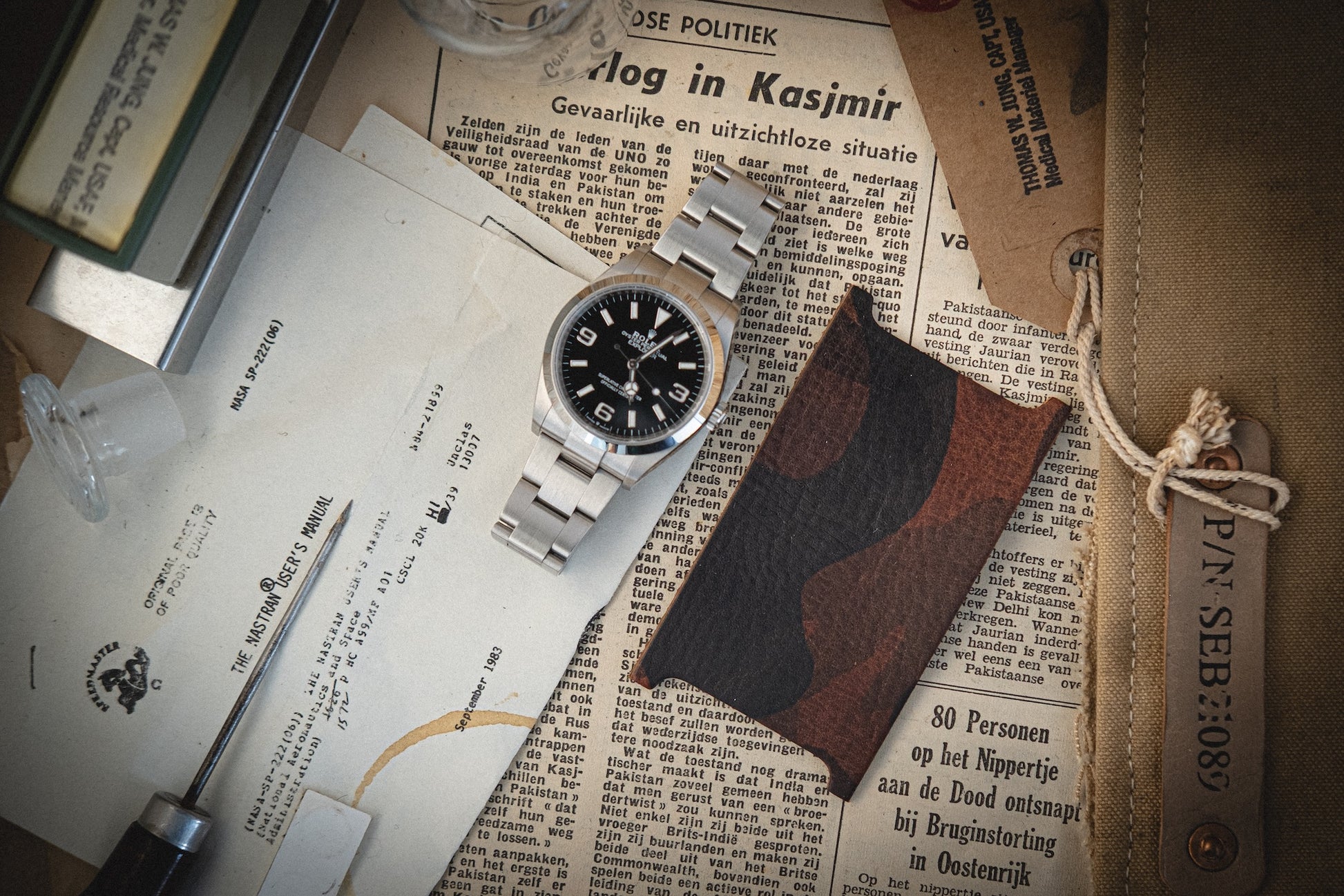 A silver wristwatch with a black dial sits on vintage papers, surrounded by a pick tool and leather tag inscribed with "PIN: SEB:308." Nearby, a cardholder from the Camo veg-tan collection in brown and black camo adds charm to the beige surface.