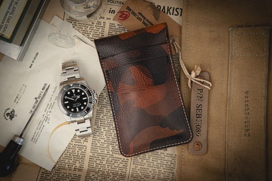 A silver-banded watch with a dark face lies on vintage newspaper clippings next to a brown camo veg-tan leather pouch, a small screwdriver, and assorted documents. A beige fabric label partially visible hints at its handmade in Antwerp quality, from the Camo veg-tan collection.