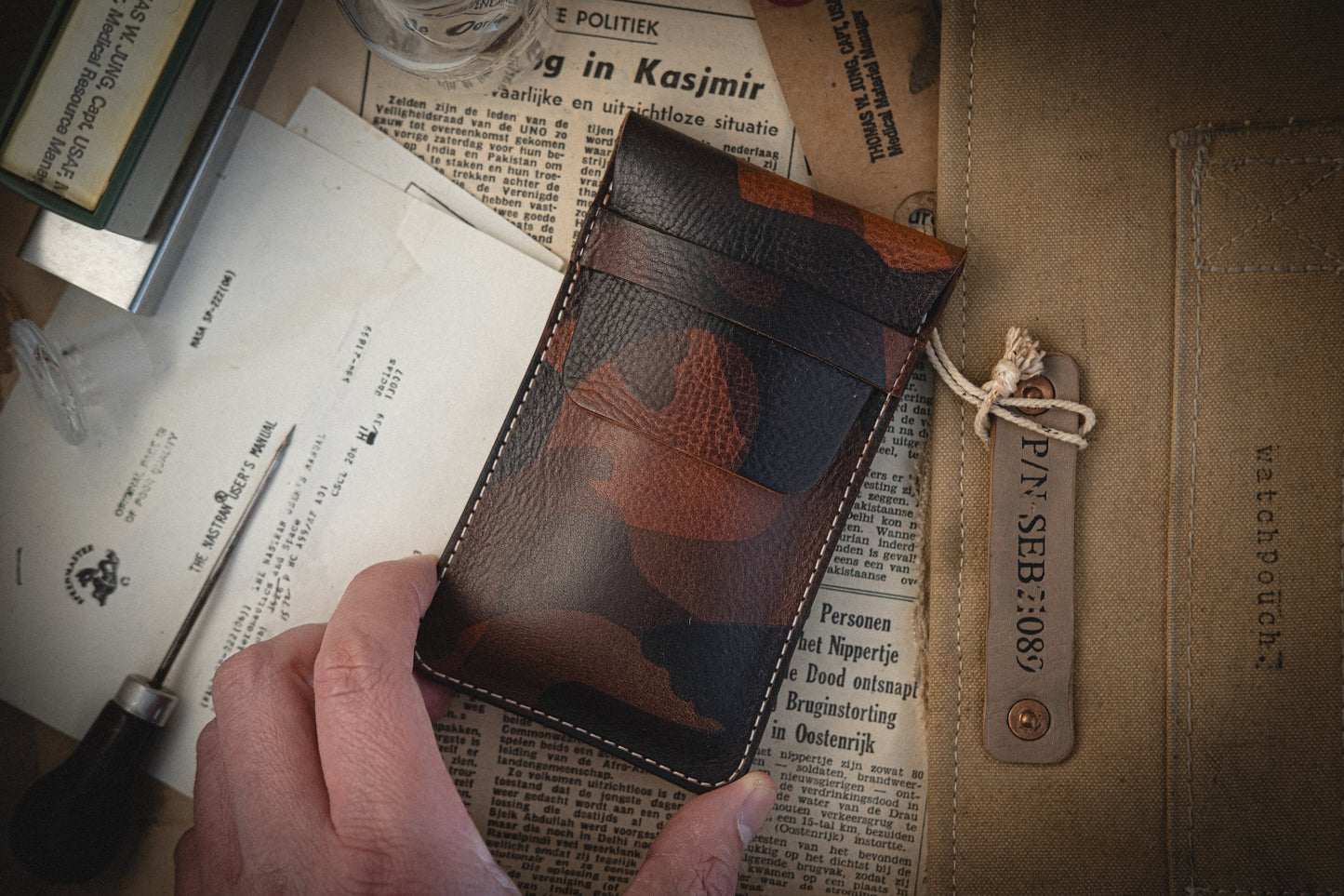 A hand holds a Camo veg-tan leather wallet above vintage newspapers. Nearby, a watch pouch embroidered with "SEB:089" lies amid writing tools. Handmade in Antwerp, these pieces offer personalization for discerning collectors.