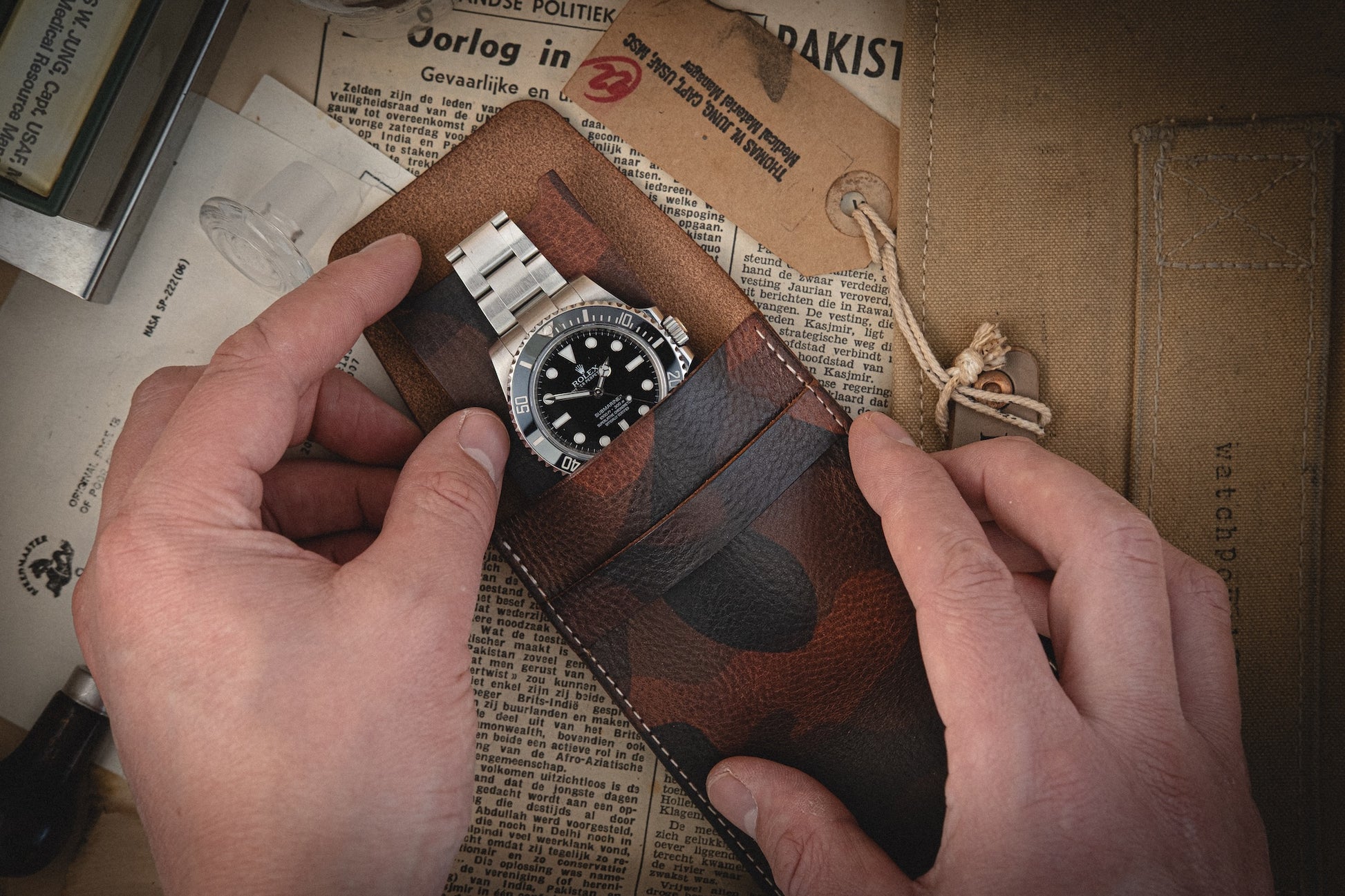 Hands holding a wristwatch with a metal strap are nestled in the Camo veg-tan collection's leather case. This handmade Antwerp watch has a black dial. Set against an old newspaper, string-tied envelope, and booklet, it captures a vintage and nostalgic vibe perfectly suited for personalized accessories.