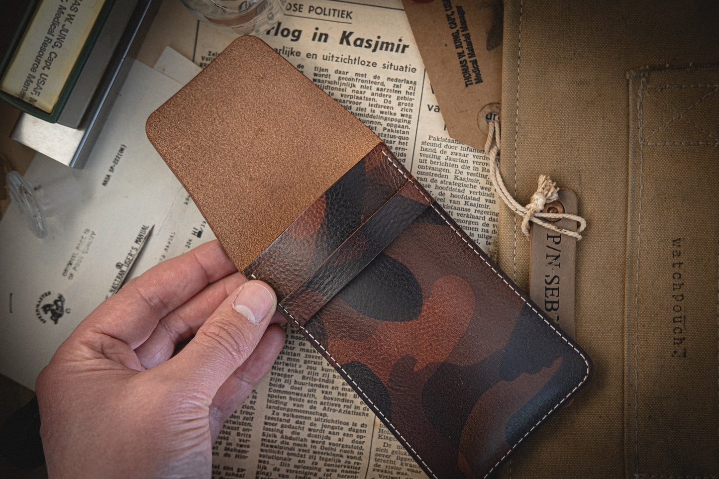 A hand showcasing an open wallet from the Camo veg-tan collection, handmade in Antwerp, rests on a table filled with papers. A partially visible paper folder with a string closure sits beside it, complementing the organized chaos.