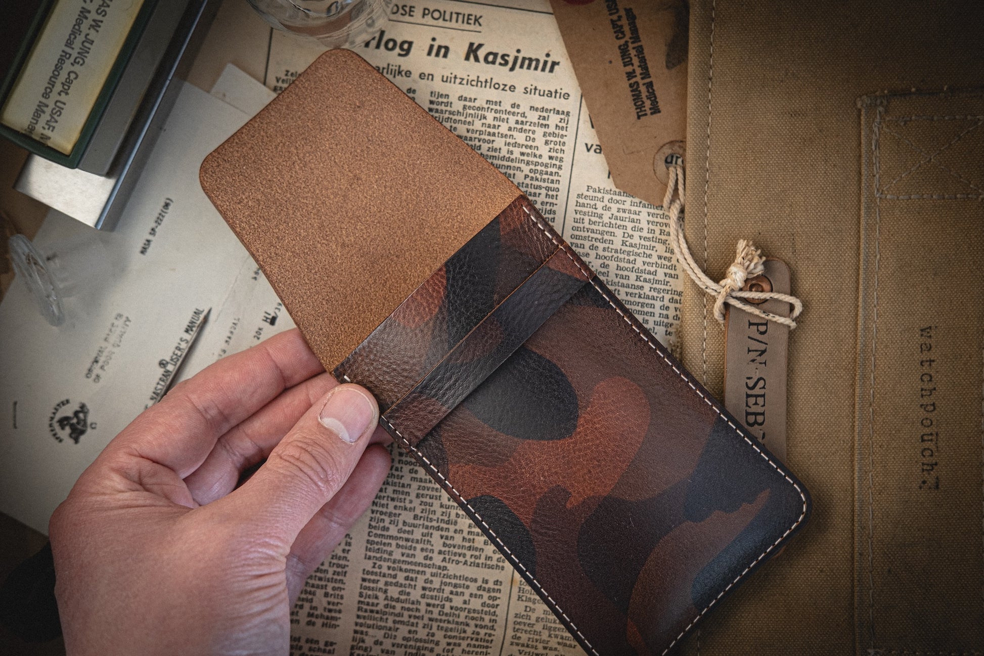 A hand showcasing an open wallet from the Camo veg-tan collection, handmade in Antwerp, rests on a table filled with papers. A partially visible paper folder with a string closure sits beside it, complementing the organized chaos.