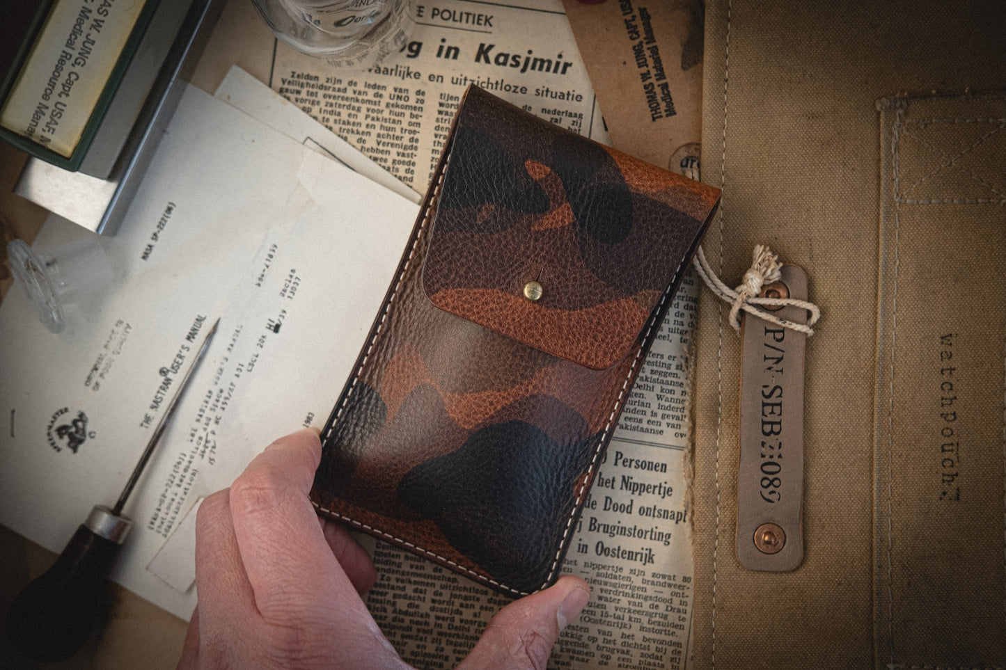 A hand holds a pouch from the Camo veg-tan collection. Below are various papers and a foreign language newspaper clipping. To the right, a canvas watch pouch with a "WATCHPOUCH" leather tag rests, all intricately handmade in Antwerp.